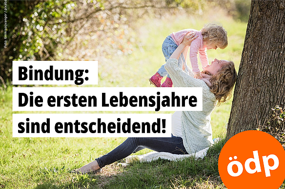 Eine Frau sitzt auf dem Boden, mit dem Rücken zu einem Baum und hält ein Kind hovh über ihren Kopf. Beide lachen.